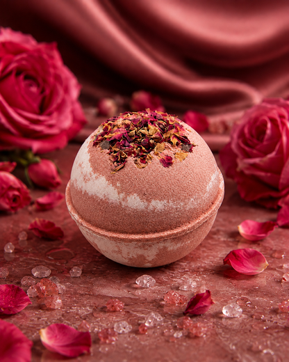 A spherical bath bomb with dusty rose and white marbled pattern,(or an uncolored option is available), scented with florals, amber, vanilla, and musk, topped with dried rose petals, set on a rustic surface surrounded by red roses, rose petals, and pink bath salts.
