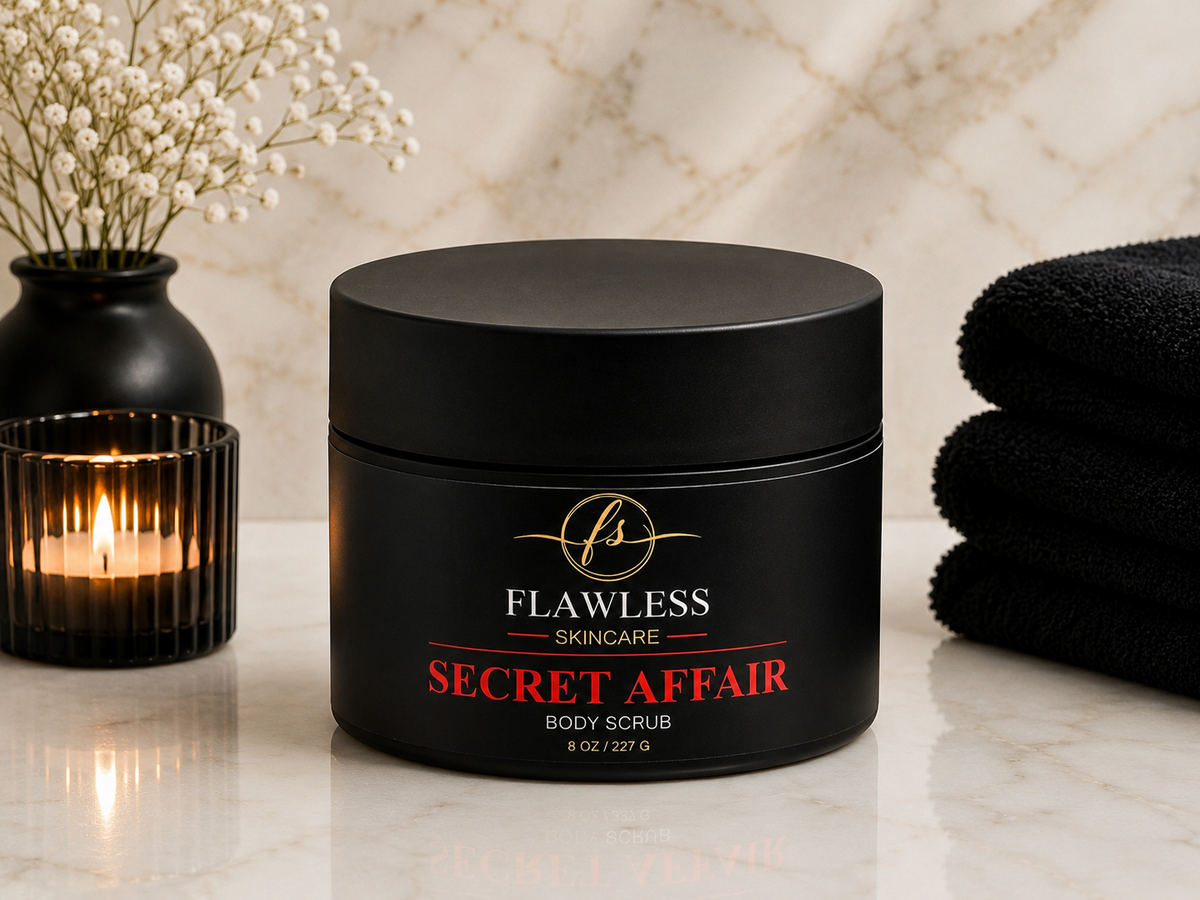 This is a man’s scented body scrub with musk, amber, citrus, and woody notes. A black jar of Flawless Skincare 'Secret Affair' body scrub sits on a marble surface, styled with a lit candle, dried flowers, and folded black towels.