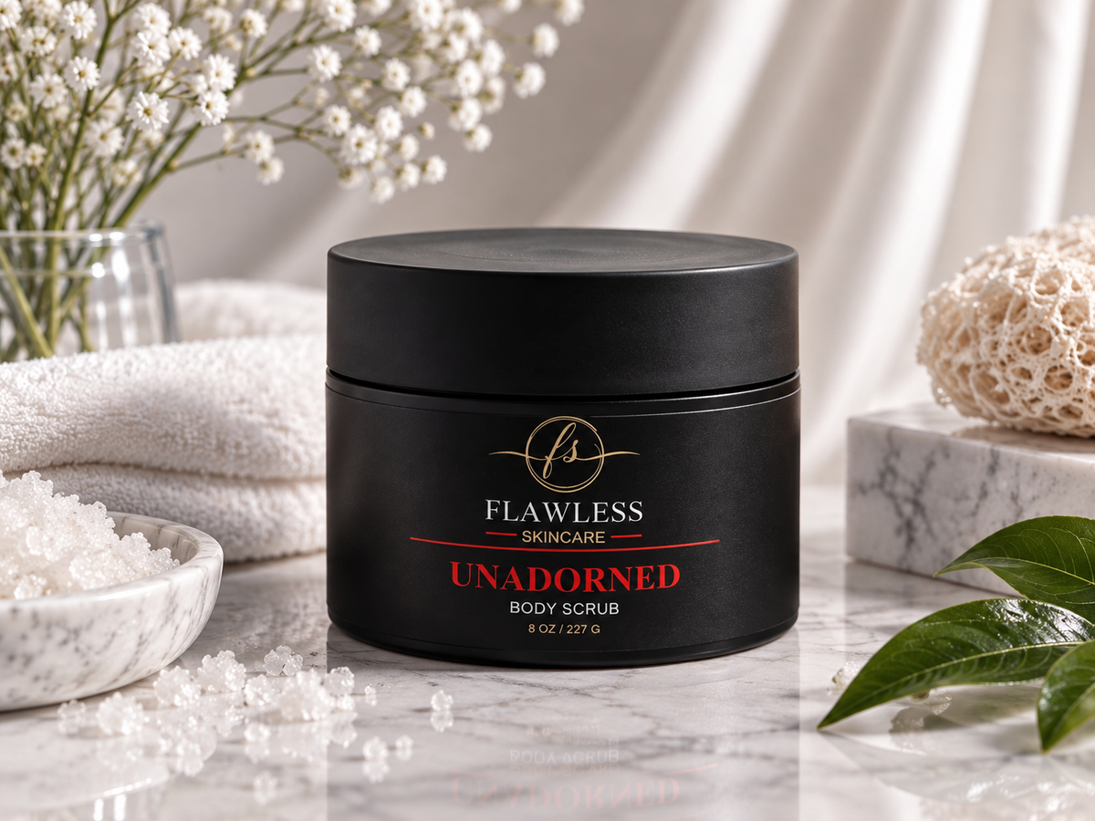 A black jar of Flawless Skincare 'Unadorned', (unscented), Body Scrub sits on a marble surface surrounded by spa props including white flowers, folded towels, a bowl of scrub, and green leaves.