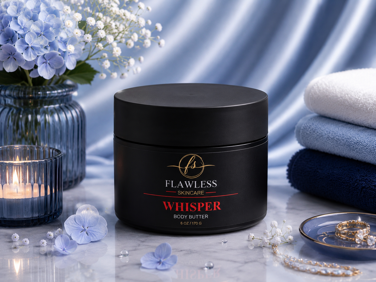 This is a scented body butter with amber and floral notes. A black jar of Flawless Skincare Whisper Body Butter with scents of warm amber and florals. It is centered on a marble surface, surrounded by blue flowers, a lit candle, folded towels, and jewelry against a blue satin backdrop.
