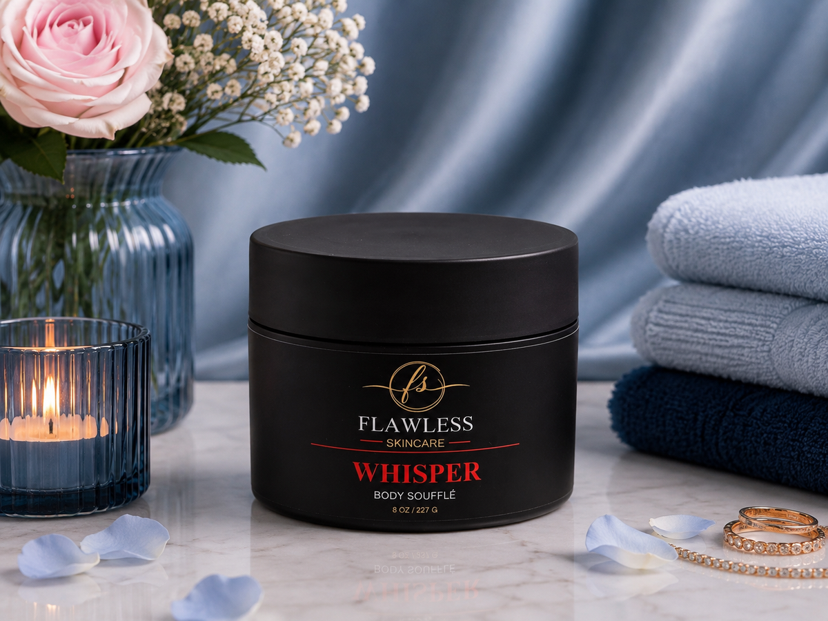 This is a scented body lotion with amber and floral notes. A black jar of Flawless Skincare 'Whisper' Body Soufflé centered on a marble surface, styled with a pink rose in a blue vase, a lit candle, folded towels, and rings against a blue satin backdrop.